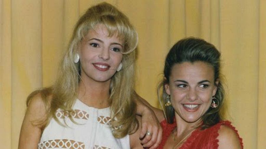 Dos mujeres sonríen juntas; una con vestido blanco y la otra con vestido rojo.  Cabello rubio y largo en una, castaño claro recogido en la otra. Fondo dorado.