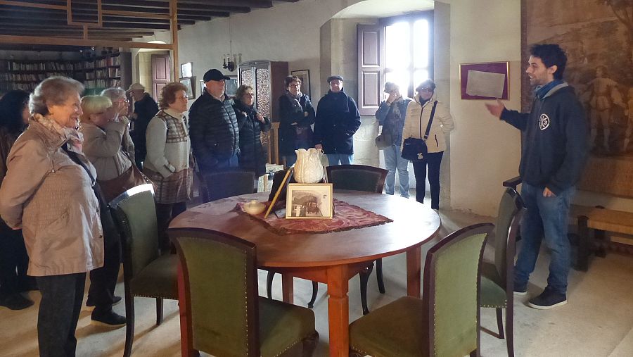 Guía turístico explica a un grupo la historia de un lugar, señalando un documento enmarcado en la pared de una sala con biblioteca.