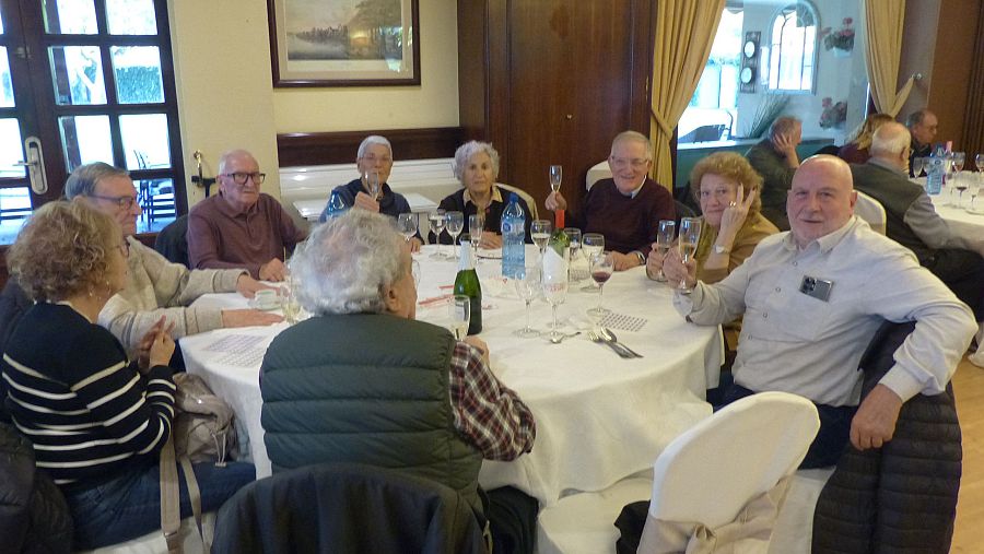 Reunión festiva de personas mayores alrededor de una mesa con bebidas, en un espacio interior con paredes de madera. Ambiente distendido.