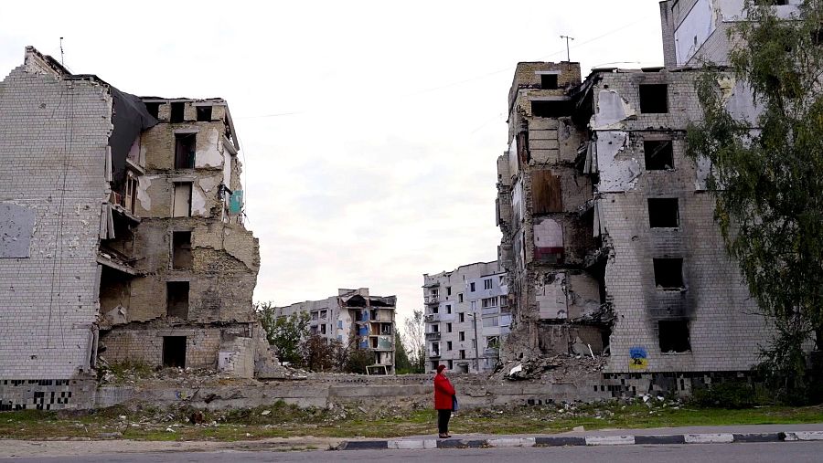 Edificios residenciales destruidos, escombros y una persona con abrigo rojo observando la escena. Daños significativos en fachadas y ventanas.