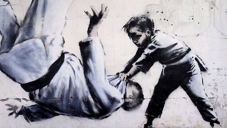 Grafiti de Banksy: David, en ropa oscura, usa judo para vencer a Goliat, en judogi blanco, en una pared deteriorada.  Metáfora de la resistencia ucraniana.