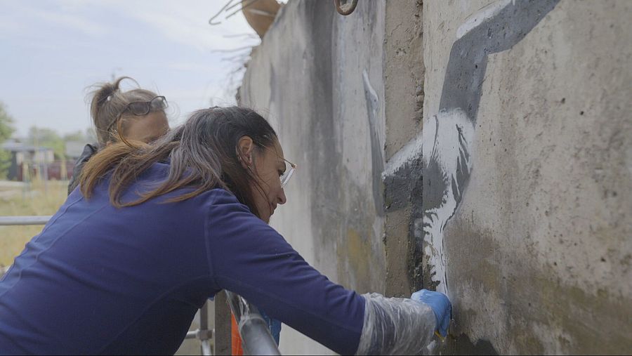 Restauradora aplica material sobre grieta en graffiti de una gimnasta, en Ucrania.  Usa guantes y camisa azul oscuro.  Otra mujer observa.