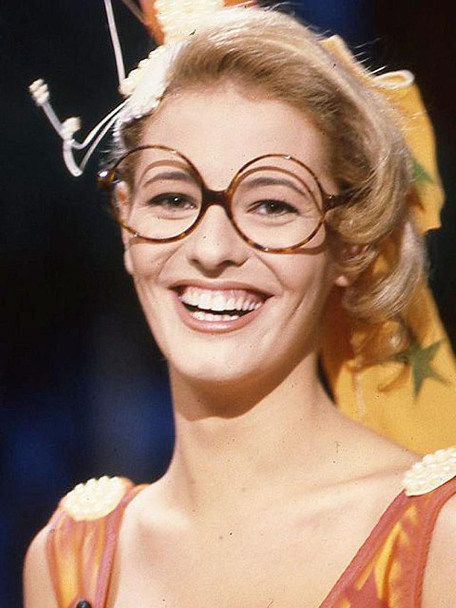 Presentadora con gafas redondas y blusa naranja y amarilla, cabello recogido y sonrisa amplia en un escenario televisivo.