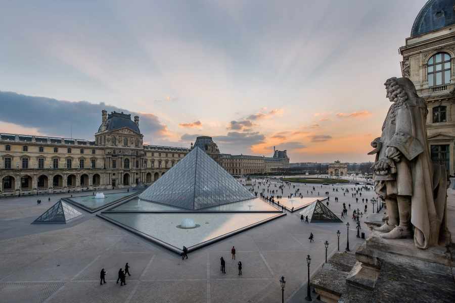 Vista panorámica del Louvre al atardecer, con su pirámide de cristal y gente paseando en la plaza. El cielo muestra tonos cálidos.