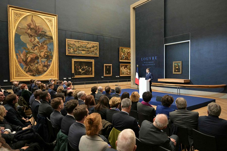 Comparecencia de Emmanuel Macron en el Louvre. El presidente francés, de pie ante un atril y la bandera francesa, se dirige a un público en una sala con pinturas y la Mona Lisa visible.