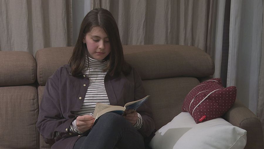Joven leyendo un libro en un sofá, con camisa oscura y jersey a rayas, cabello castaño y expresión concentrada. Ambiente tranquilo.