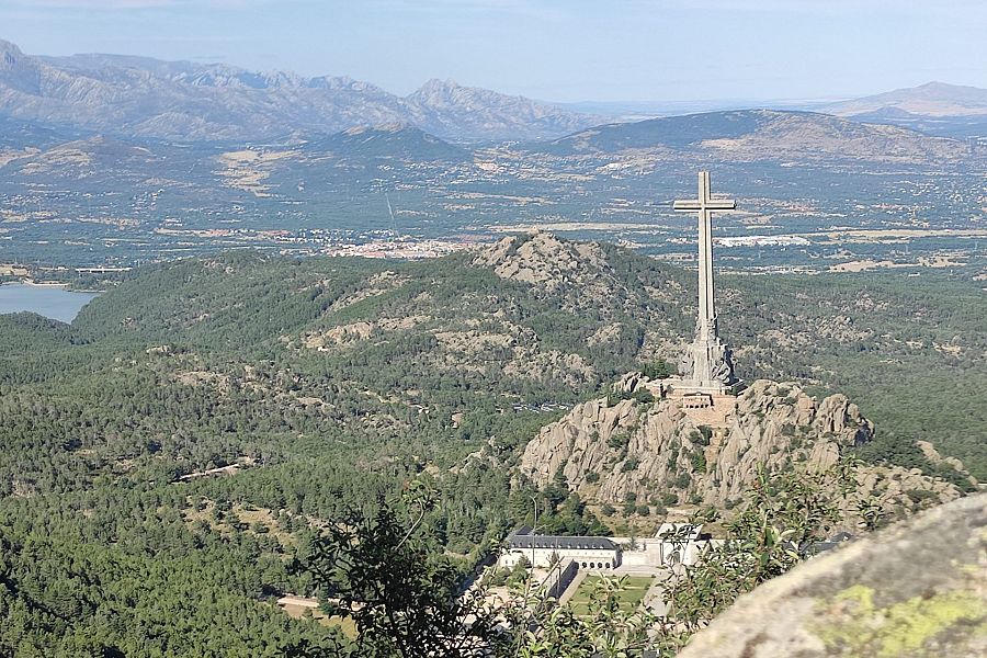 Impresionante vista panorámica de una gran cruz en una montaña rocosa, rodeada de abundante vegetación y un cuerpo de agua.