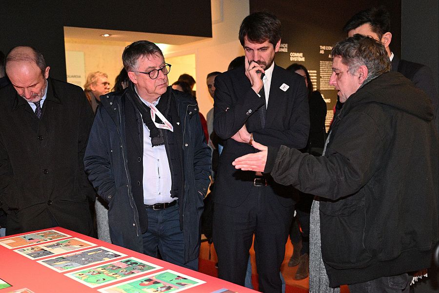 Ministro de Cultura escuchando atentamente a un hombre que le muestra ilustraciones de cómics en una mesa con mantel rojo.  Ambiente de galería con otras personas observando.