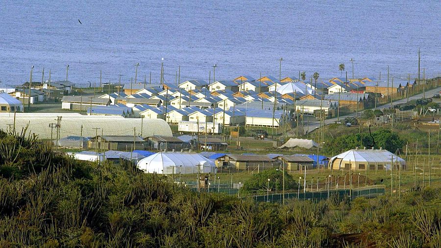 Vista aérea de una base naval con edificios, vegetación y el mar al fondo. Se aprecian estructuras de diversos tamaños y materiales.