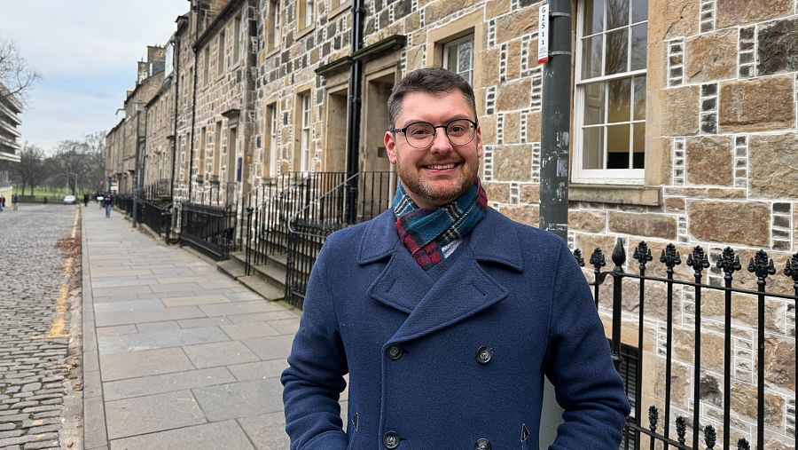 Profesor de mediana edad con abrigo azul, bufanda a cuadros y camisa blanca, sonriendo a la cámara en una calle con edificios de piedra y zona verde al fondo.
