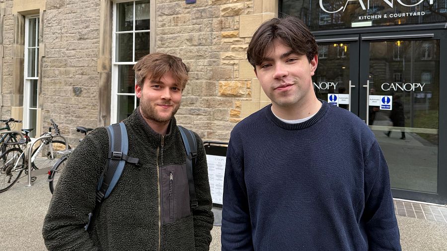Dos jóvenes sonríen a la cámara frente a un restaurante de piedra con grandes ventanales y el nombre 