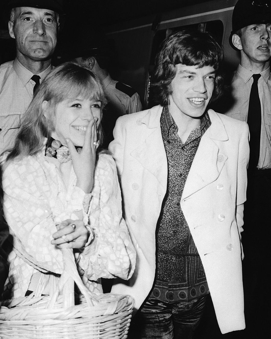 Imagen en blanco y negro de Marianne Faithfull y Mick Jagger en una estación de tren. Ella lleva una blusa estampada y una cesta; él, una chaqueta blanca.  Policías de uniforme al fondo.