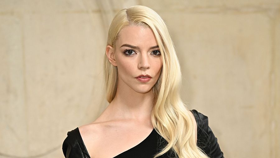 Retrato de Anya Taylor-Joy con vestido negro asimétrico, cabello rubio platino y mirada seria a cámara.