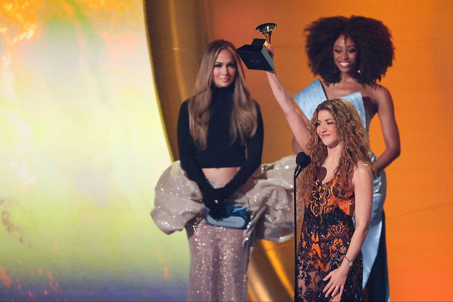 Shakira recibe un Grammy, acompañada de Jennifer Lopez y otra mujer.  Shakira luce un vestido semitransparente y cabello rizado; Jennifer Lopez, un top negro y falda plateada.