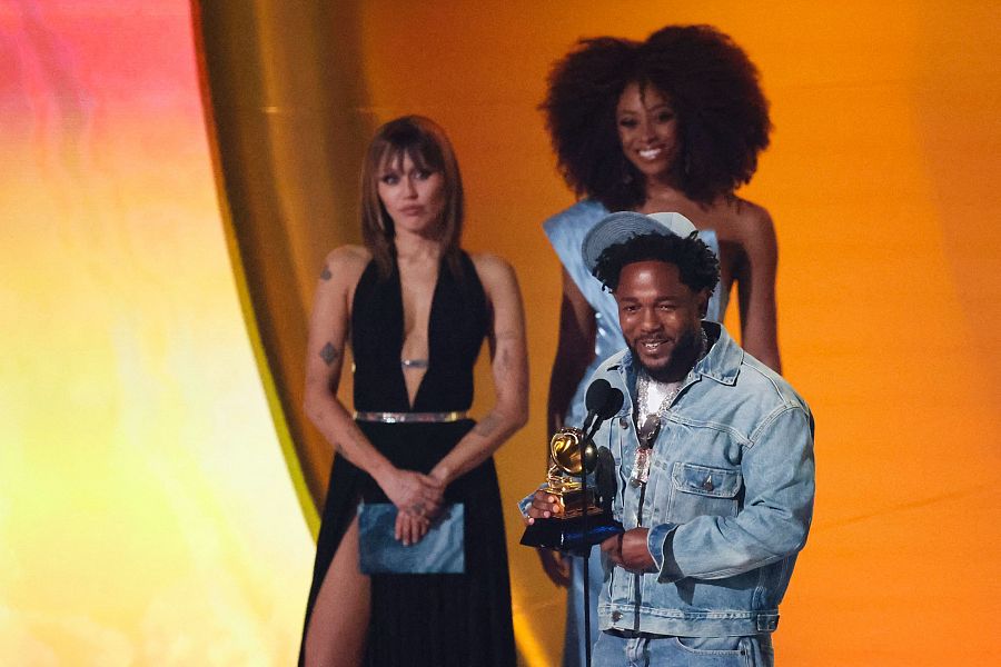 Kendrick Lamar, con atuendo informal, sonríe al recibir un Grammy.  Acompañado por Miley Cyrus y otra mujer.