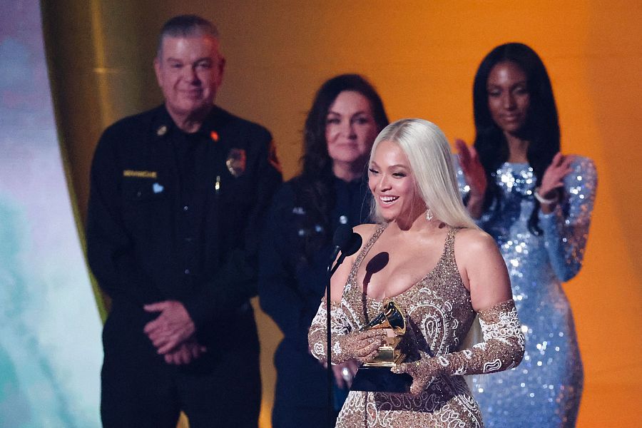 Beyoncé, radiante en un vestido dorado, sostiene el gramófono del Grammy al Álbum del Año.  A su alrededor, otras personas la felicitan.