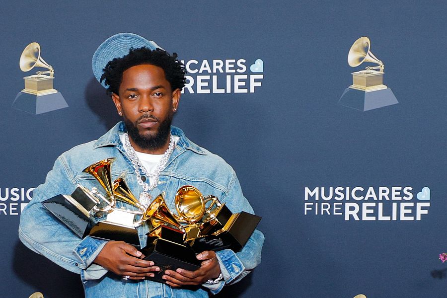 Rapero con chaqueta y gorro vaqueros sosteniendo cuatro premios Grammy en evento benéfico. Fondo azul oscuro con texto 