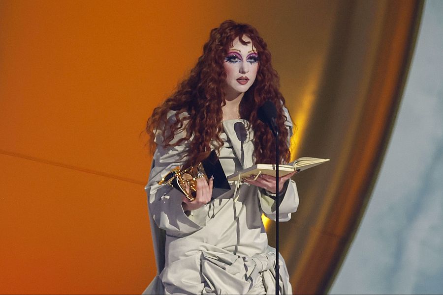 Artista con pelo rojo y maquillaje intenso recibe un premio, leyendo aparentemente un discurso desde una libreta.