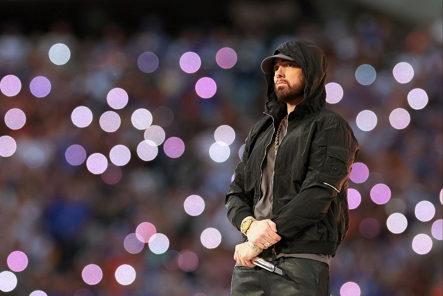 Eminem en el escenario, con chaqueta y gorra negras, rapea ante una multitud desenfocada con luces de fondo creando un efecto bokeh.