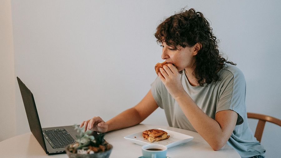 Mujer con pelo rizado comiendo un bollo mientras trabaja en un portátil;  a su alrededor, una taza, un plato con más bollos y una planta.