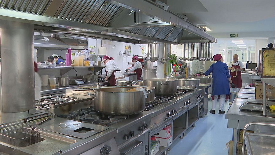 Cocineros uniformados preparan comida en una cocina industrial española, usando grandes ollas y equipos profesionales.