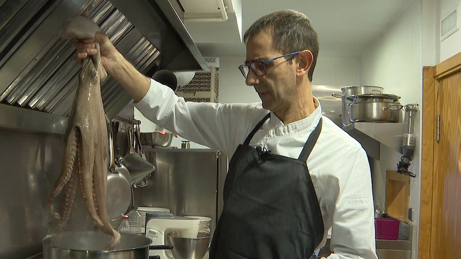Chef con gafas sujetando un pulpo sobre una olla hirviendo en una cocina profesional.  Preparación culinaria de alta cocina.