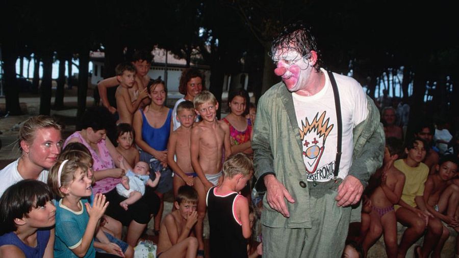 Payaso con traje verde oliva sobre camiseta blanca actuando ante niños y mujeres en un espacio abierto.  Momento de alegría en medio de un conflicto bélico.