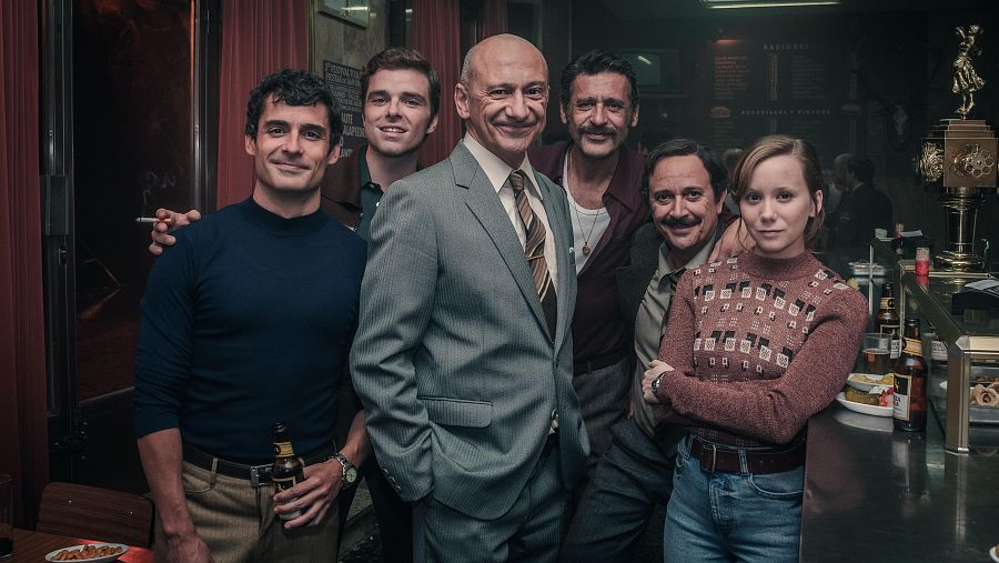 Seis actores de 'Asuntos Internos' en un bar, con atuendos variados: jerseys, camisas, trajes. Ambiente informal, estética de época.