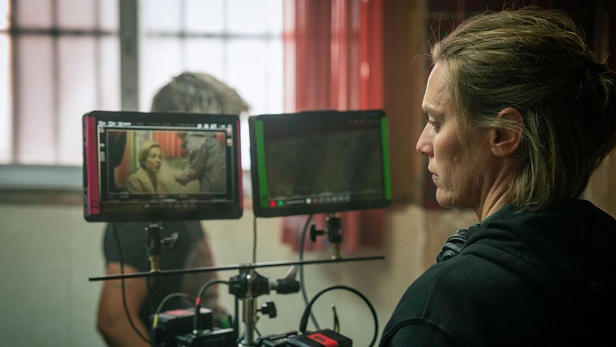 Detrás de cámaras: mujer del equipo de rodaje observa dos monitores mostrando una escena con dos mujeres en una habitación. Cables y soportes visibles.