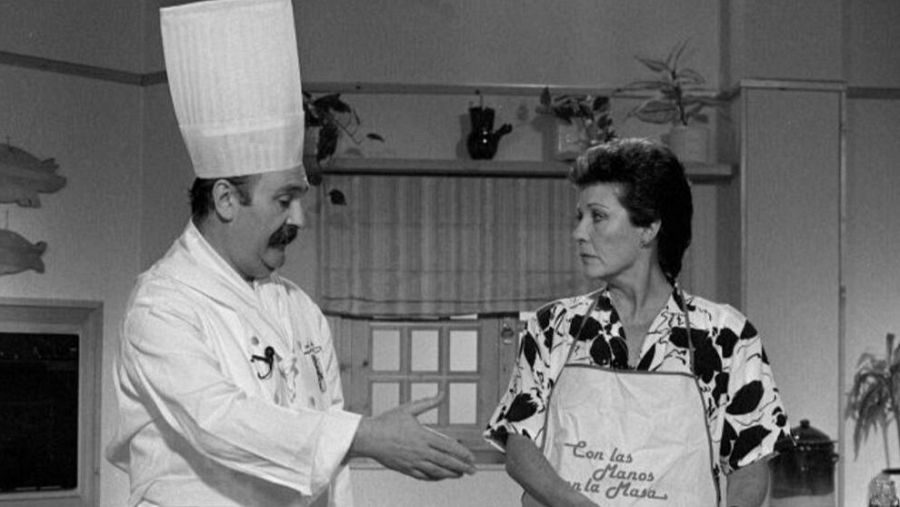 Escena de cocina en blanco y negro: un chef instruye a una presentadora con delantal. Decoración con siluetas de peces.