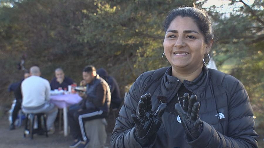 Mujer sonriente con chaqueta negra y guantes, en un espacio abierto con personas al fondo sentadas a una mesa.