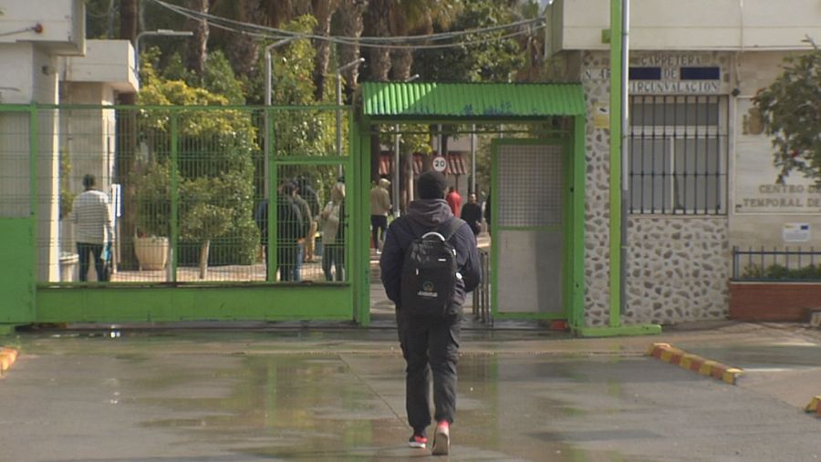 Hombre con mochila negra y zapatillas rosas se aleja de una entrada con verja verde; suelo mojado; personas visibles tras la verja.
