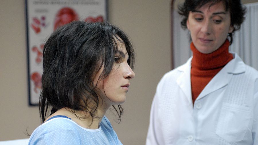 Dos mujeres, una paciente con bata azul y una doctora con bata blanca y jersey naranja, se miran con seriedad en una consulta médica. Póster anatómico en la pared.