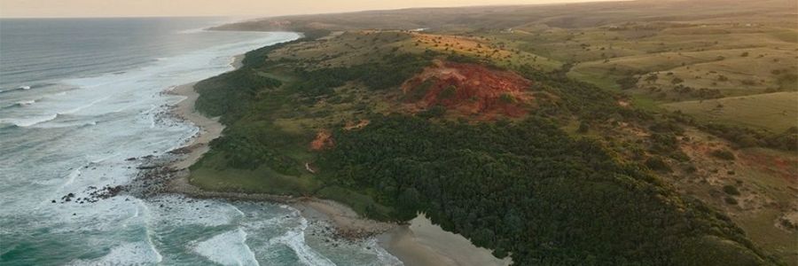 Vista aérea de una costa rocosa con océano azul, vegetación verde oscura y una colina rojiza al atardecer.  Topografía variada.