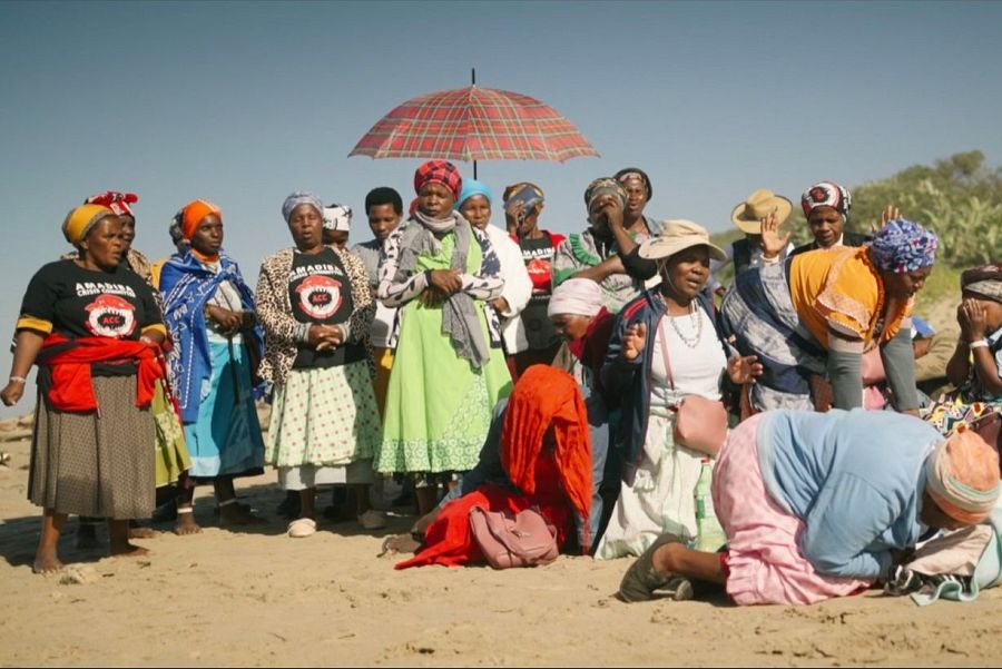 Ritual en playa sudafricana: mujeres AmaMpondo, vestidas con atuendos tradicionales y camisetas, realizan una ceremonia con expresiones de concentración y devoción junto al mar.