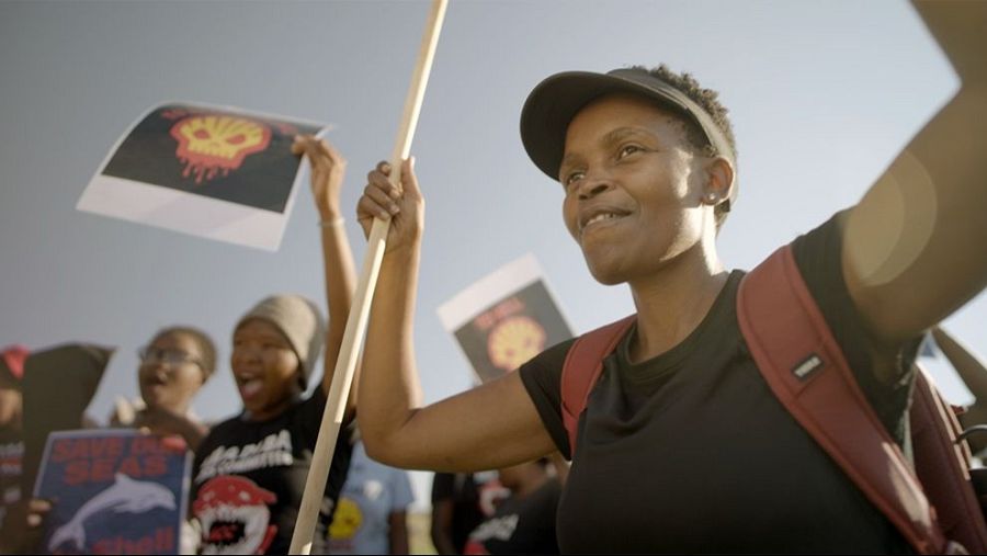 Activista con gorra y mochila roja en manifestación, sosteniendo un palo largo.  Se observan pancartas con mensajes ambientales y una calavera con una gota de petróleo.