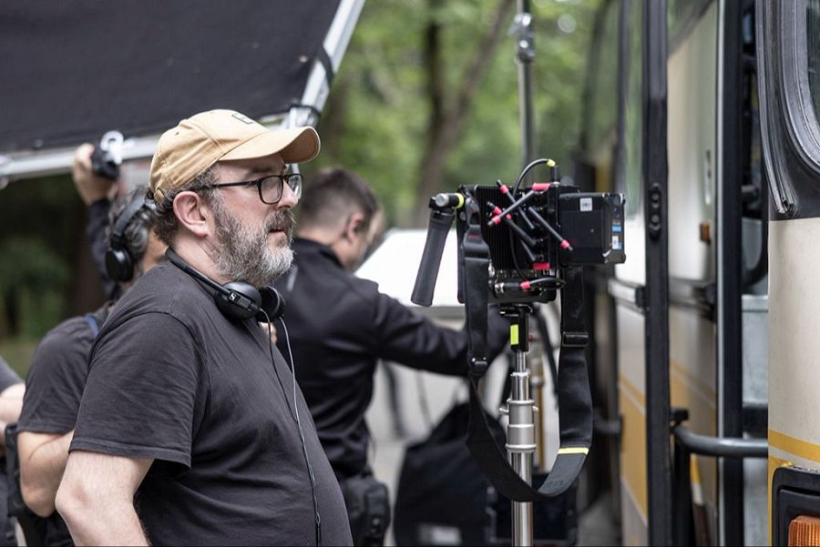 Borja Cobeaga, con gorra y auriculares, supervisa una escena en un rodaje exterior.  Un miembro del equipo, también con auriculares, está presente. Se ve parte de un autobús y equipo de filmación.