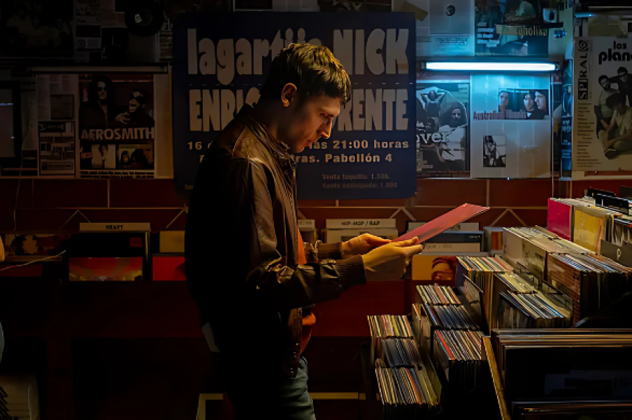 Hombre examina un disco en tienda de música con carteles de conciertos y vinilos. Se aprecian fragmentos de texto en los carteles: 