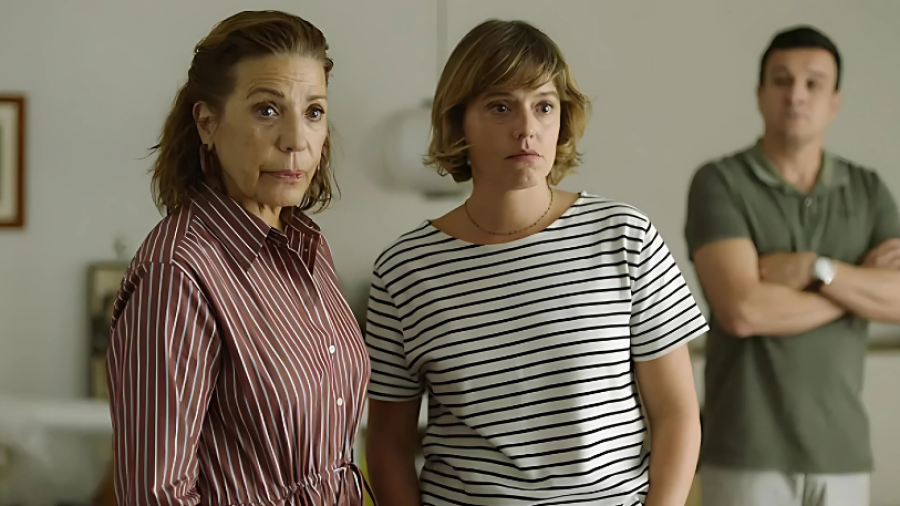 Dos actrices, con camisas a rayas y expresiones serias, conversan en primer plano. Un hombre con camiseta verde observa desde el fondo.