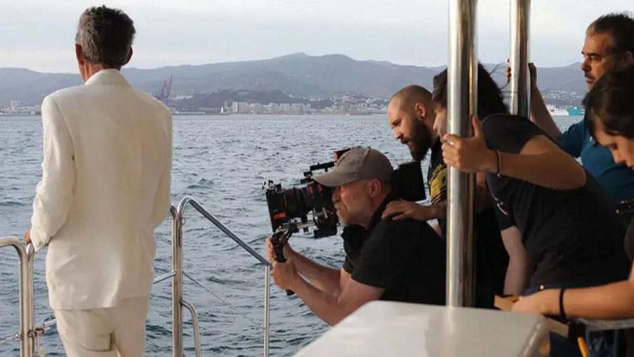 Escena de rodaje en barco con un hombre de traje blanco como protagonista, equipo de filmación trabajando y ciudad costera al fondo.
