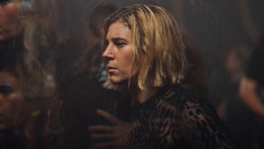 Primer plano de mujer con cabello rubio corto y blusa oscura, expresión seria.  Fotograma de película con fondo oscuro y borroso.