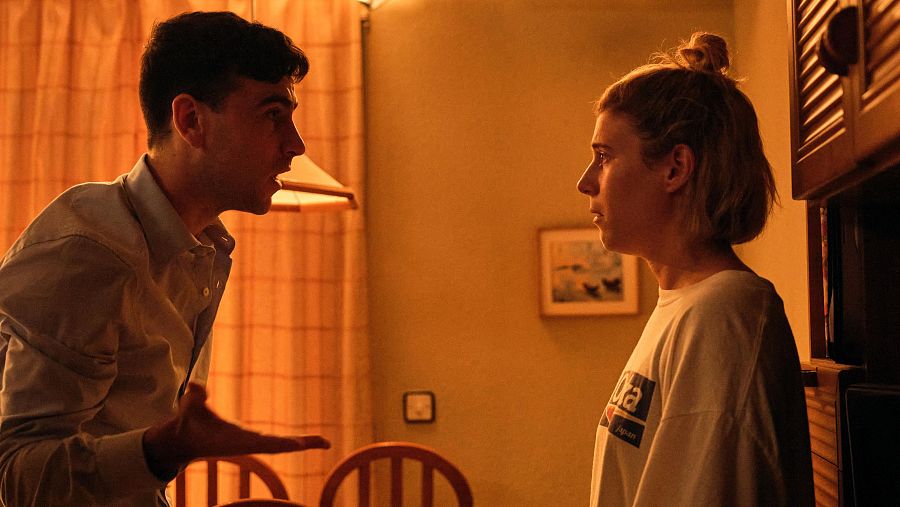 Escena de discusión entre un hombre y una mujer en un interior doméstico. Él gesticula con enojo; ella, seria, observa. Tonos cálidos y ambiente tenso.