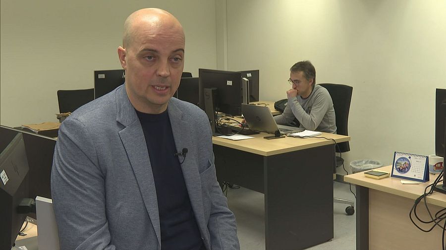 Primer plano de un hombre con americana gris y camiseta negra hablando a cámara en una oficina con otros empleados y ordenadores.