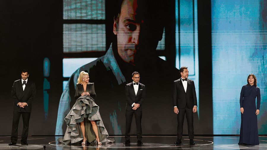 Cinco personas en elegante atuendo participan en un homenaje en los Premios Goya.  Una gran pantalla muestra una imagen de fondo.