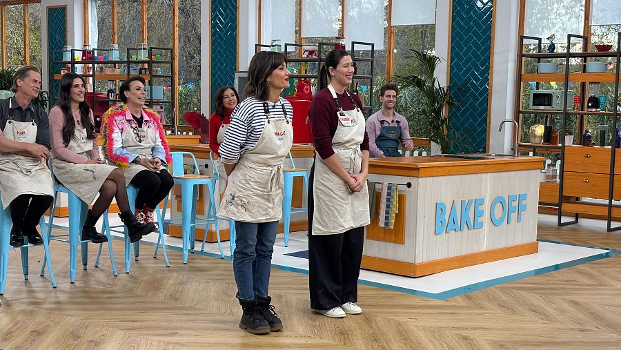 Dos mujeres con delantales en el plató de Bake Off, junto a un mostrador.  Ambiente luminoso con participantes al fondo.