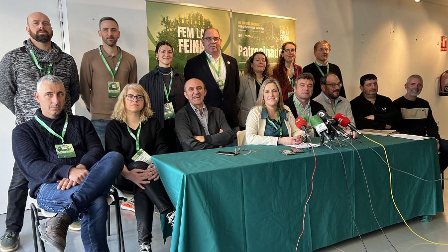 Grupo de 14 personas, hombres y mujeres, posan tras una mesa con micrófonos.  Llevan credenciales de Unió de Pagesos.  Se ven carteles con el logo del sindicato.