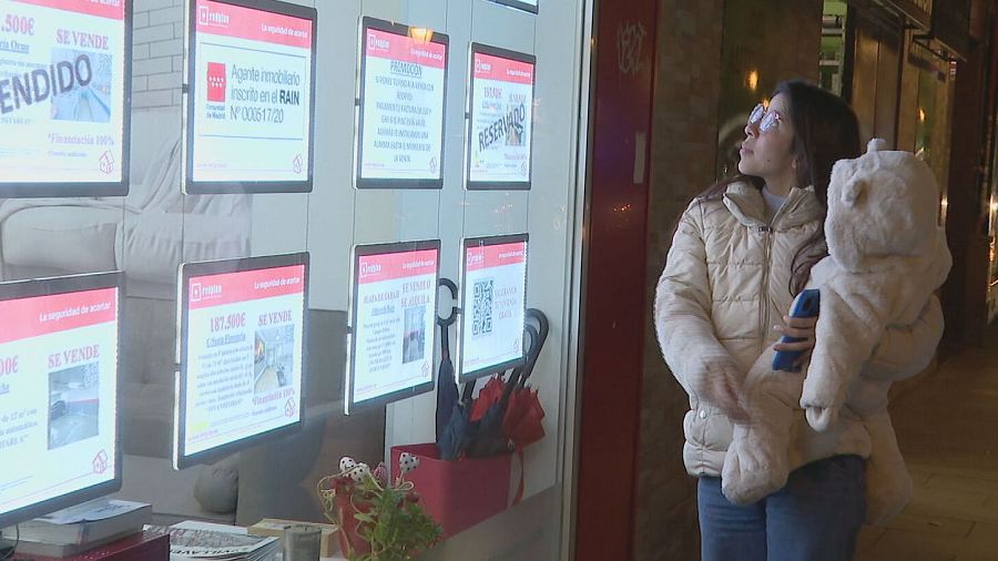 Mujer con bebé observa anuncios inmobiliarios con precios y carteles de 