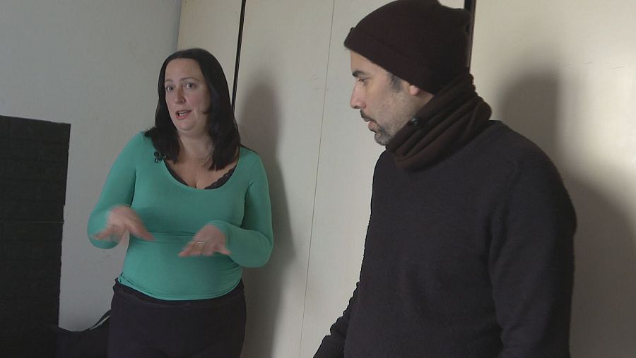 Dos personas conversan en una habitación sencilla. Una mujer con camiseta verde gesticula; el hombre, con gorro y bufanda marrones, la escucha.