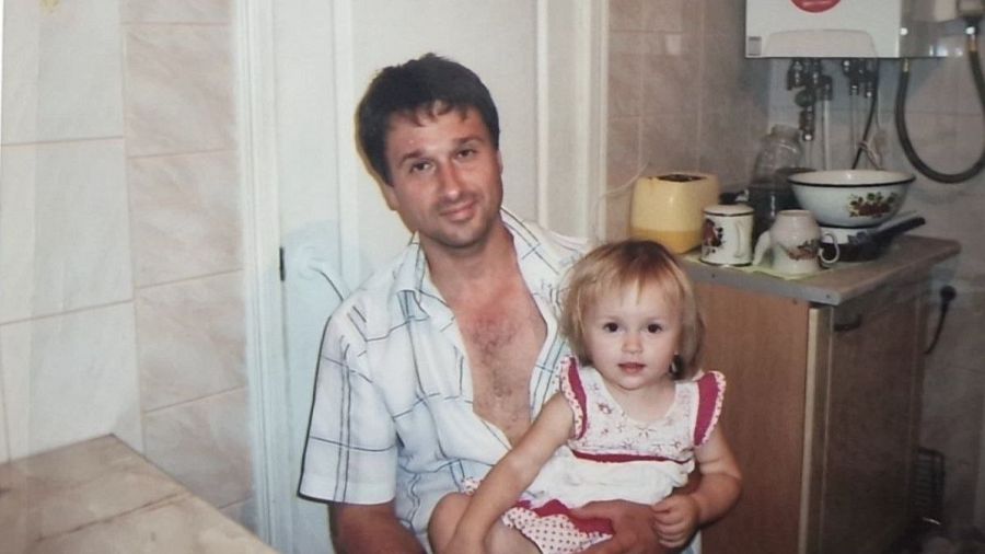 Hombre y niña en una cocina o baño con azulejos blancos. El hombre lleva una camisa a cuadros y la niña un vestido blanco con detalles rosas.  Hay utensilios de cocina visibles.
