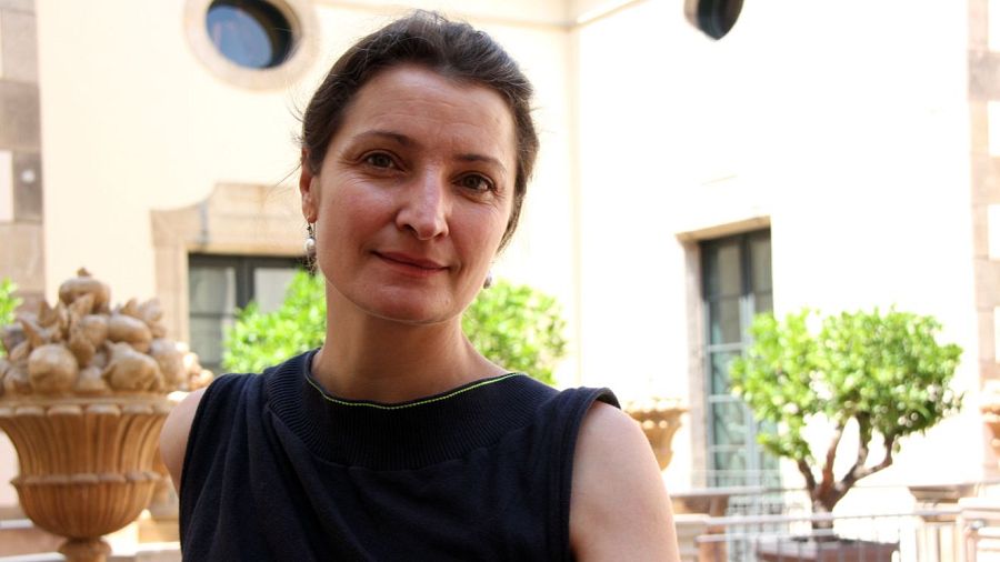 Retrato de Catherine Allard con blusa negra y pendientes, en un patio con arcos y macetas.  Aparece la palabra 